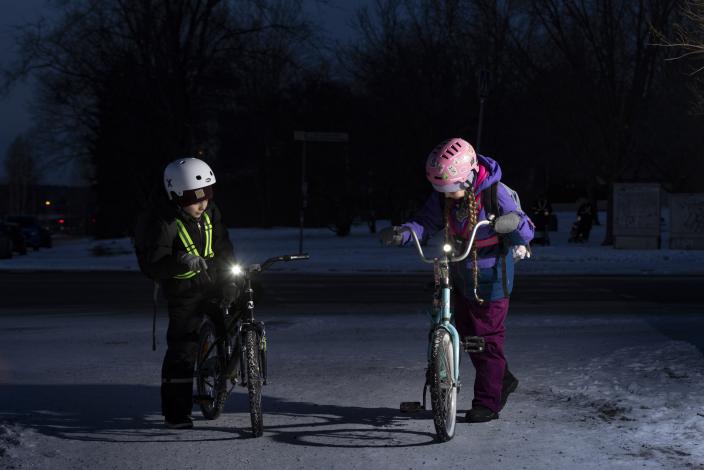 Kaksi lasta heijastimilla ja kypärät päässä seisovat pyöriensä vieressä pimeässä ja lumisessa illassa. (Kuva: Suvi Elo)