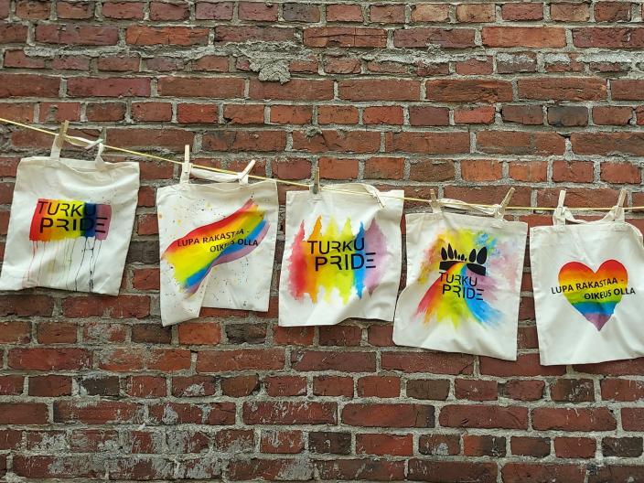 Kuvassa roikkuu pyykkinarulla viisi Pride-aiheista kangaskassia. Taustalla on tiiliseinä.
