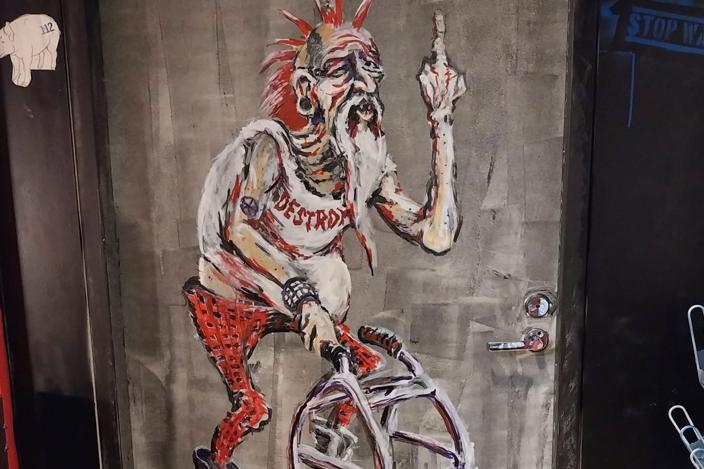 Kuvassa seinään maalattu iäkäs punkkari, joka kulkee rollaattorilla ja näyttää keskisormea.