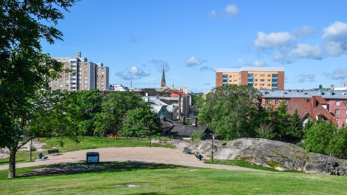 Maisema Puolalanpuistosta sataman suuntaan. Puistoalueella nurmikkoa, hiekka-aukio, jonka reunoilla kaksi penkkiä ja kalliota. Taustalla kerrostaloja ja Mikaelinkirkon torni. (Kuva: Henri Taponen)