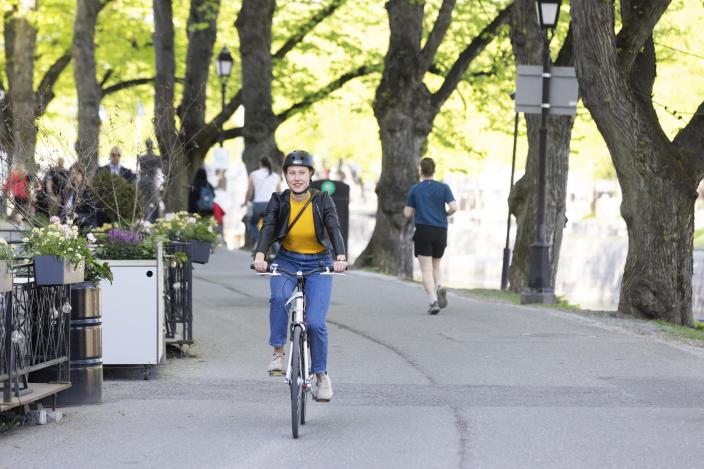 Nainen pyöräilee Turun jokirannassa kesällä, taustalla näkyy puita ja muita ihmisiä. (Kuva: Suvi Elo)