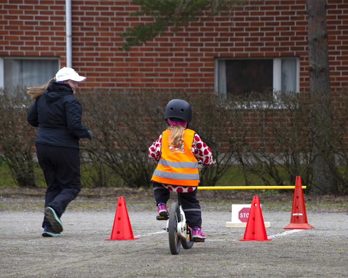 Lapsi pyöräilee ulkona pyöräilytaitoradalla, aikuinen juoksee vieressä. (Kuva: Jalmari Salaterä)