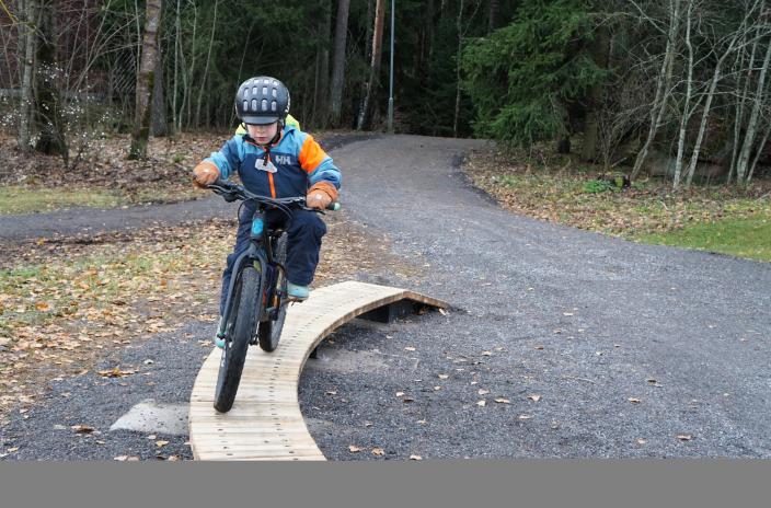 Lapsi pyöräilee pyörärataelementtinä olevan rampin päältä, taustalla näkyy metsää ja polkuja. (Kuva: Matti Koistinen)