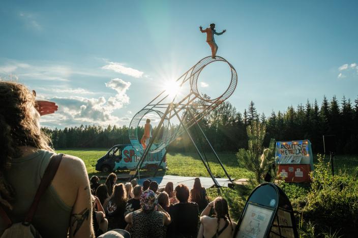 Yleisö seuraa kesäisessä puistossa esitystä, jossa sirkustaiteilijat tasapainottelevat isokokoisessa pyörivässä rakennelmassa. (Kuva:  Nalle Elmgren)