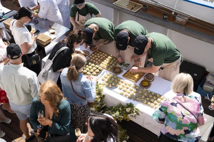 Kolme vihreäpaitaista kokkia valmistelee pieniä ruoka-annoksia ihmisille Suomen Joutsenen kannella.