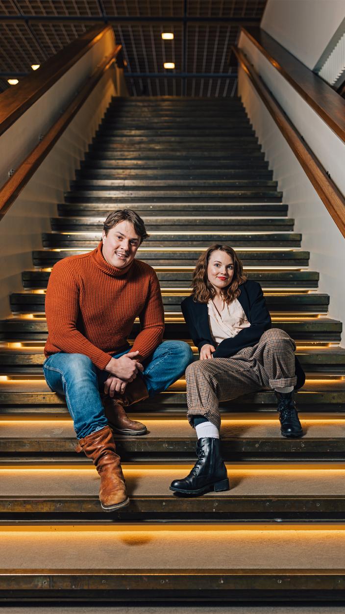  Kaksi henkilöä istuu rennosti vierekkäin isossa portaikossa teatterin lämpiössä, molemmat katsovat kameraan hymyillen. (Kuva: Heikki Räisänen)