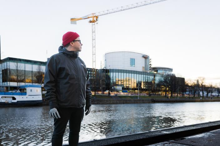 Henkilö seisoo tummiin ulkovaatteisiin ja punaiseen pipoon pukeutuneena joen rannalla ja katsoo vasemmalle. Henkilön takana vastarannalla näkyy rakenteilla oleva musiikkitalo, jonka työmaalla on myös iso nosturi. (Kuva: Linda Svarfvar)