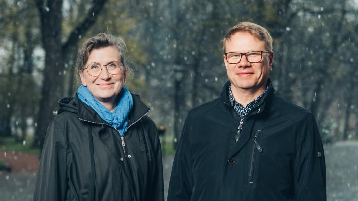 Kaksi iloisesti kameraan katsovaa henkilöä seisoo vierekkäin ulkotilassa, taustalla puistomaisema. (Kuva: Jussi Virkkumaa)