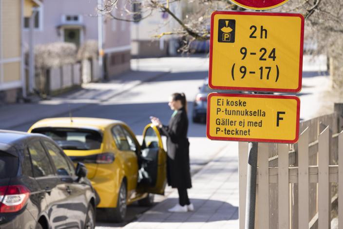 Kuvassa näkyy kadulla oleva pysäköintikyltti, taustalla näkyy katu, jonka varrella nainen katsoo puhelintaan auton vierellä.  (Foto: Suvi Elo)