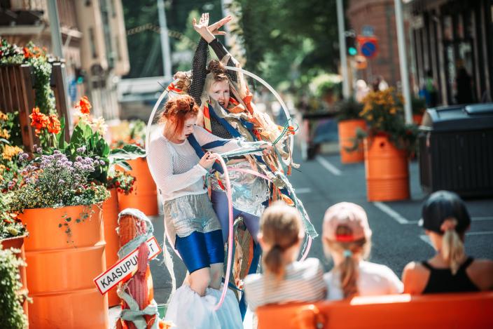 Lapset katsovat Turun kesäkadulla katuteatteriesitystä. Lasten edessä esiintyy kaksi henkilöä, joista toinen yrittää pujottautua hulavanteen läpi. (Kuva: Jussi Virkkumaa)