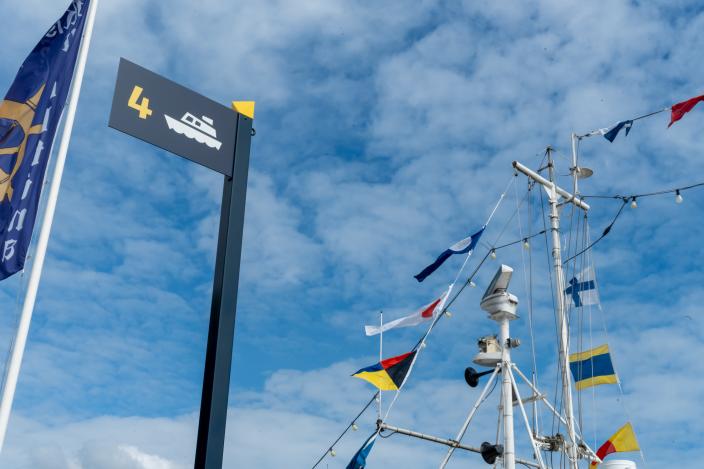 En svart skylt med en bild av ett fartyg och siffran fyra. Till höger finns en mast med flaggor fästa på. I bakgrunden syns en blå himmel och moln. (Foto: Focus-Video)