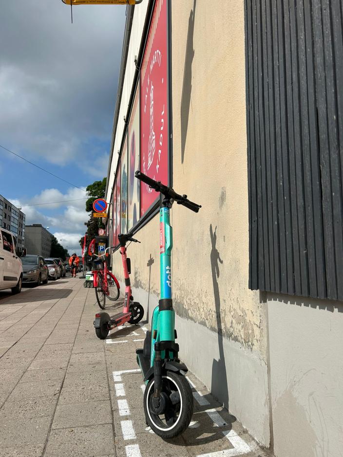 Sähköpotkulauta siististi pysäköitynä kadulla, jossa näkyy skuuttiparkki. (Kuva: Lilli Louhikoski)