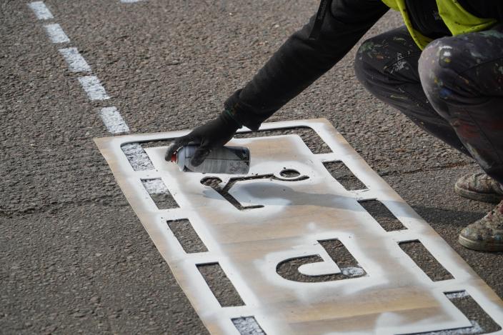 Läheltä otettu kuva henkilöstä maalaamassa spray-maalilla sähköpotkulautojen parkkiruutuja. Henkilöllä on apunaan ruutujen maalaamiseen tarkoitettu sapluuna. (Kuva: Iiris Yli-Junnila)