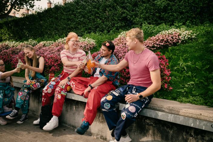 Viisi kaverusta jokirannassa opiskelijahaalarit päällä, neljä istuu reunuksella ja yksi istuu pyörätuolissa. Kilistelevät limupulloja ja hymyilevät. (Kuva: Antti Merisaari / Mama Creative)
