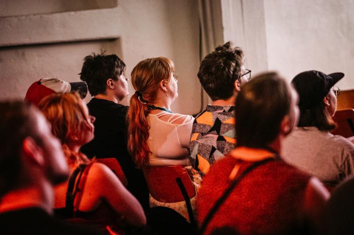 Elokuvasalin sisältä otettu kuva viistosti yleisön takaa. Yleisö katsoo kuvan ulkopuolelle jäävää valkokangasta, joka valaisee salissa istuvien henkilöiden ääriviivat. (Kuva: Jussi Virkkumaa)