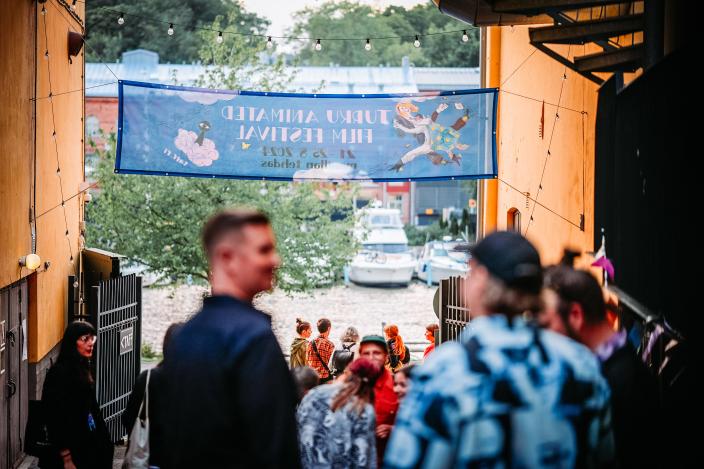 Ulkona päiväsaikaan otettu kuva, jonka edustalla ihmisiä Manillan kulttuuritehtaan sisäpihalla. Taustalla näkyvät TAFFin banderolli ja Aurajoen maisema. (Kuva: Jussi Virkkumaa)