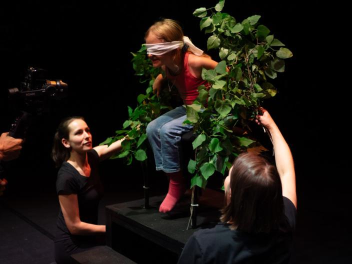 Silmälapulla varustettu lapsi osallistuu näyttämöllä esitykseen vihreiden oksien ympäröimänä kahden esiintyjän avustamana. (Kuva: Eye Dance International)