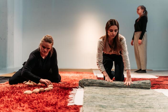 Kolme näyttelijää, joista yksi tekee kivistä jonoa, yksi rullaa mattoa ja yksi seisoo taustalla,. (Kuva: Jussi Virkkumaa)