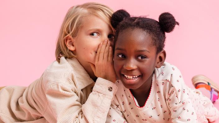 Two children are lying on their stomachs on the floor, leaning on their arms. One of them is whispering something in the other's ear. (Picture: Jenni Virta)