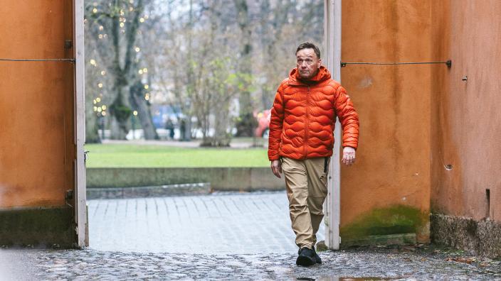 Henkilö kävelee portista kapealle kävelykadulle, jota reunustavat rakennusten ruskeanoranssit seinät. Taustalla näkyy osa aukiota puistopuineen. (Kuva: Heikki Räisänen)