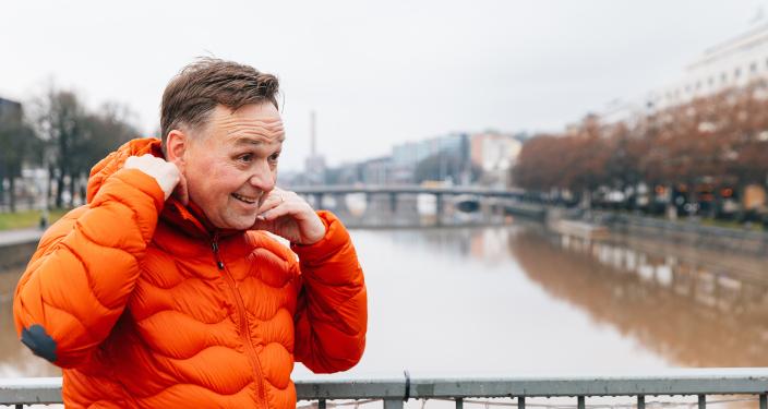 Henkilö seisoo joen ylittävällä sillalla. Hän asettelee oranssin talvitakkinsa huppua paremmin ja hymyilee samalla. Taustalla näkyy joki ja sen reunoilla puita ja rakennuksia. (Kuva: Heikki Räisänen)