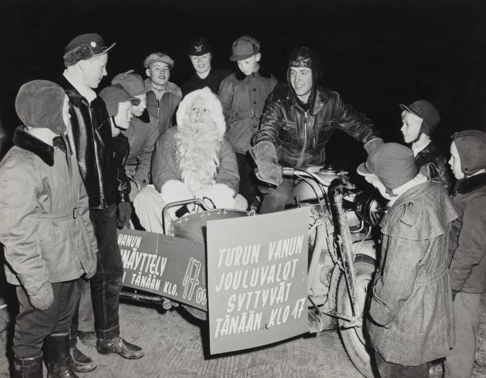 Vanha valokuva jossa moottoripyörällä istuva joulupukki on lasten ympäröimänä. (Kuva: Turun kaupunginmuseon valokuvakokoelmat)
