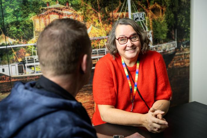 Kaksi henkilöä istuu pöydän ääressä. Toinen henkilöistä on selkä kameraan päin. Hän on kääntynyt kohti toista henkilöä, joka katsoo takaisin hymyillen. (Kuva: Jussi Vierimaa)