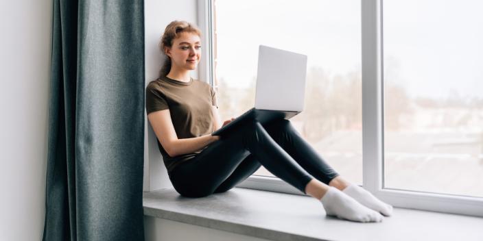 Nuori nainen istuu ikkunalaudalla ja selaa työpaikkailmoituksia kannettavalla tietokoneella. (Kuva: F8/Adobe Stock)