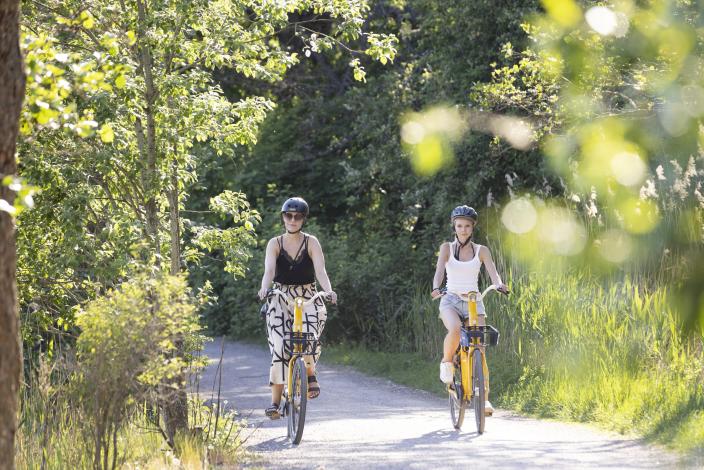 Kaksi naista pyöräilee hiekkatiellä aurinkoisena kesäpäivänä, taustalla näkyy vihreää. (Kuva: Suvi Elo)
