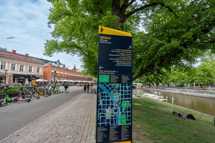 Vähätorista kertova opastekyltti, jossa on kartta ja tekstiä. Taustalla on vasemmalla Vähätori, ja oikealla kesäistä nurmikkoa, puita ja Aurajoki. (Kuva: Focus-Video)
