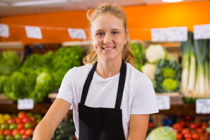 hymyilevä, mustaessuinen nuori nainen katsoo suoraan kameraan, taustalla ruokakaupan vihannestiski, jossa runsaasti vihreitä vihanneksia ja tomaatteja.